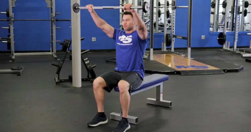Vận động viên ngồi thực hiện bài tập vai Seated Barbell Overhead Front Raise, nâng tạ đòn thẳng ra phía trước để kích hoạt mạnh cơ vai trước.