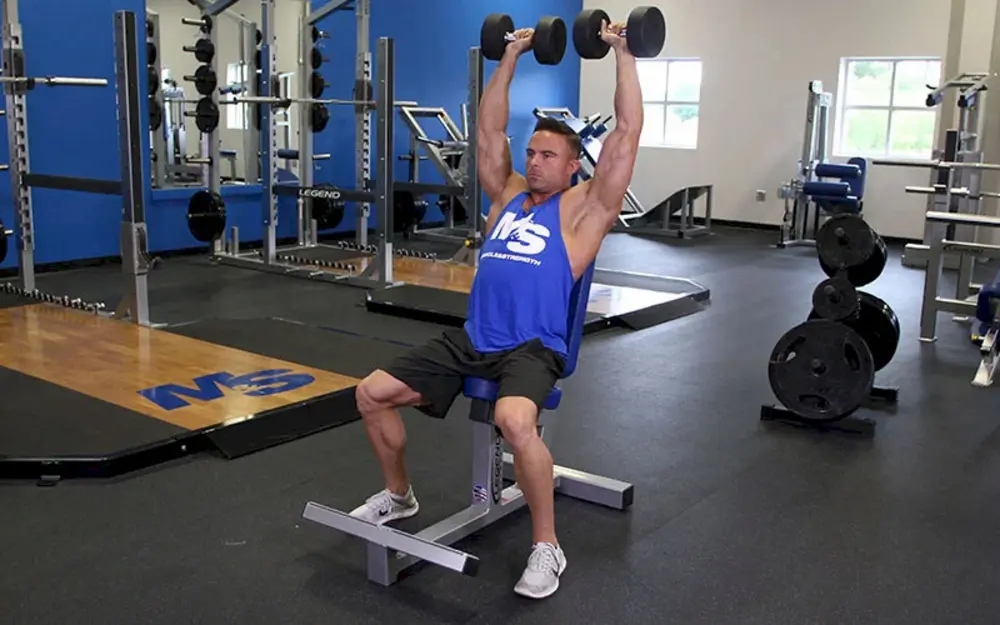Nam vận động viên tập vai Seated Dumbbell Press, ngồi trên ghế có lưng tựa và đẩy tạ đơn lên cao, giúp cô lập cơ vai và giảm áp lực lên lưng dưới.