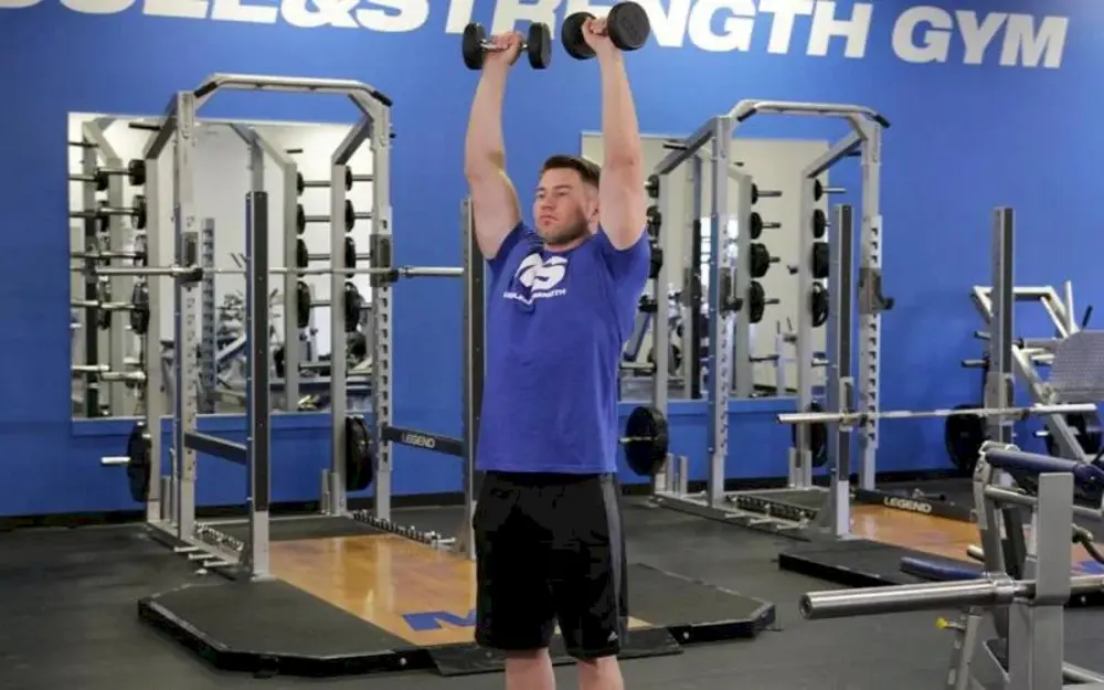 Vận động viên đứng thực hiện bài tập vai Standing Dumbbell Shoulder Press, đẩy tạ đơn qua đầu để phát triển sức mạnh toàn diện cho nhóm cơ vai và cơ core.