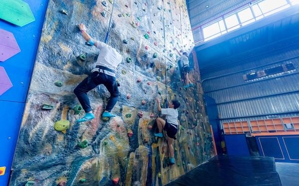 Một huấn luyện viên đang hướng dẫn một người phụ nữ cách leo núi nhân tạo, nhấn mạnh tầm quan trọng của sự hướng dẫn chuyên nghiệp