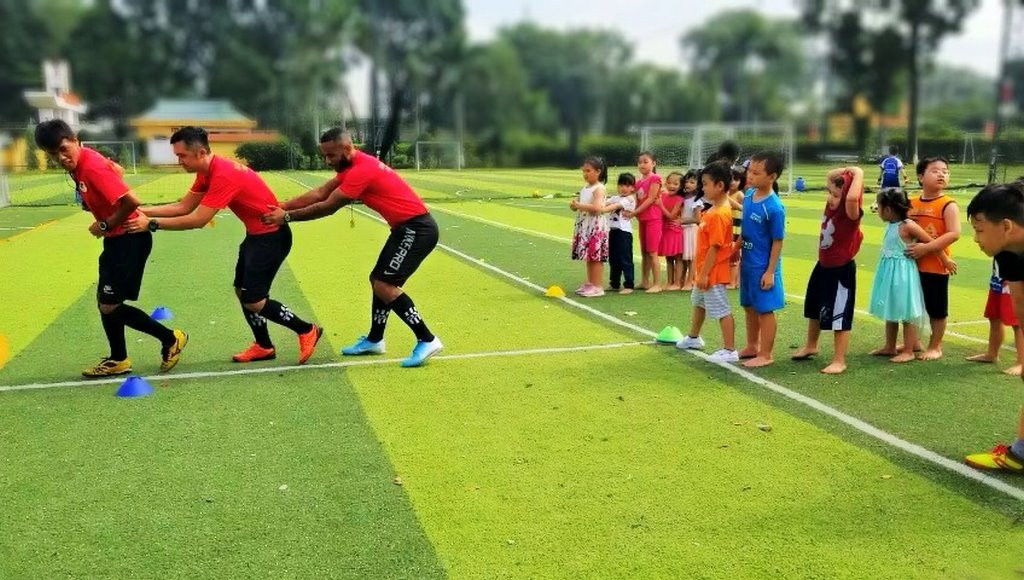Huấn luyện viên VJSS đang giảng bài trên sân cỏ nhân tạo, với bảng chiến thuật và các học viên ngồi xung quanh lắng nghe