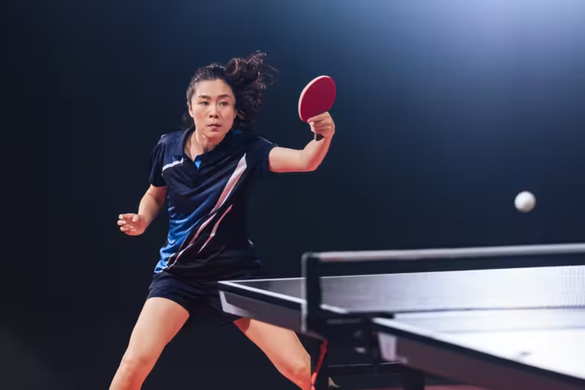 Một người phụ nữ trẻ đang đứng cạnh bàn bóng bàn, mặc áo thun thể thao và quần short, thể hiện sự thoải mái và linh hoạt cho người mới chơi bóng bàn