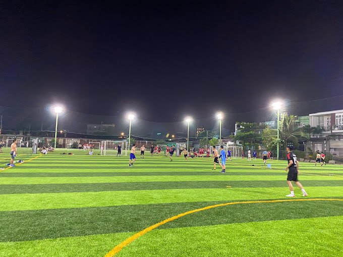 Sân bóng đá Cá Sấu Hoa Cà với cỏ nhân tạo xanh mướt, bao quanh bởi hàng rào và cây xanh, có người chơi trên sân